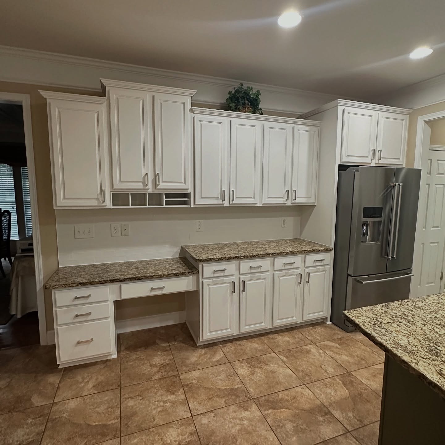 Desk and fridge wall in the finished kitchen with creamy white cabinets, silver bar pulls, cream and brown granite counters, stainless French door fridge, and warm porcelain floor tile.