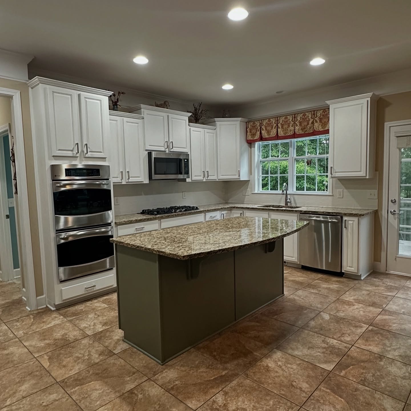 Wide view of the same kitchen now with creamy white perimeter cabinets, a sage green island with a granite top, the same cream and brown granite counters, same warm porcelain floor tile, and the same red paisley valance over the window.