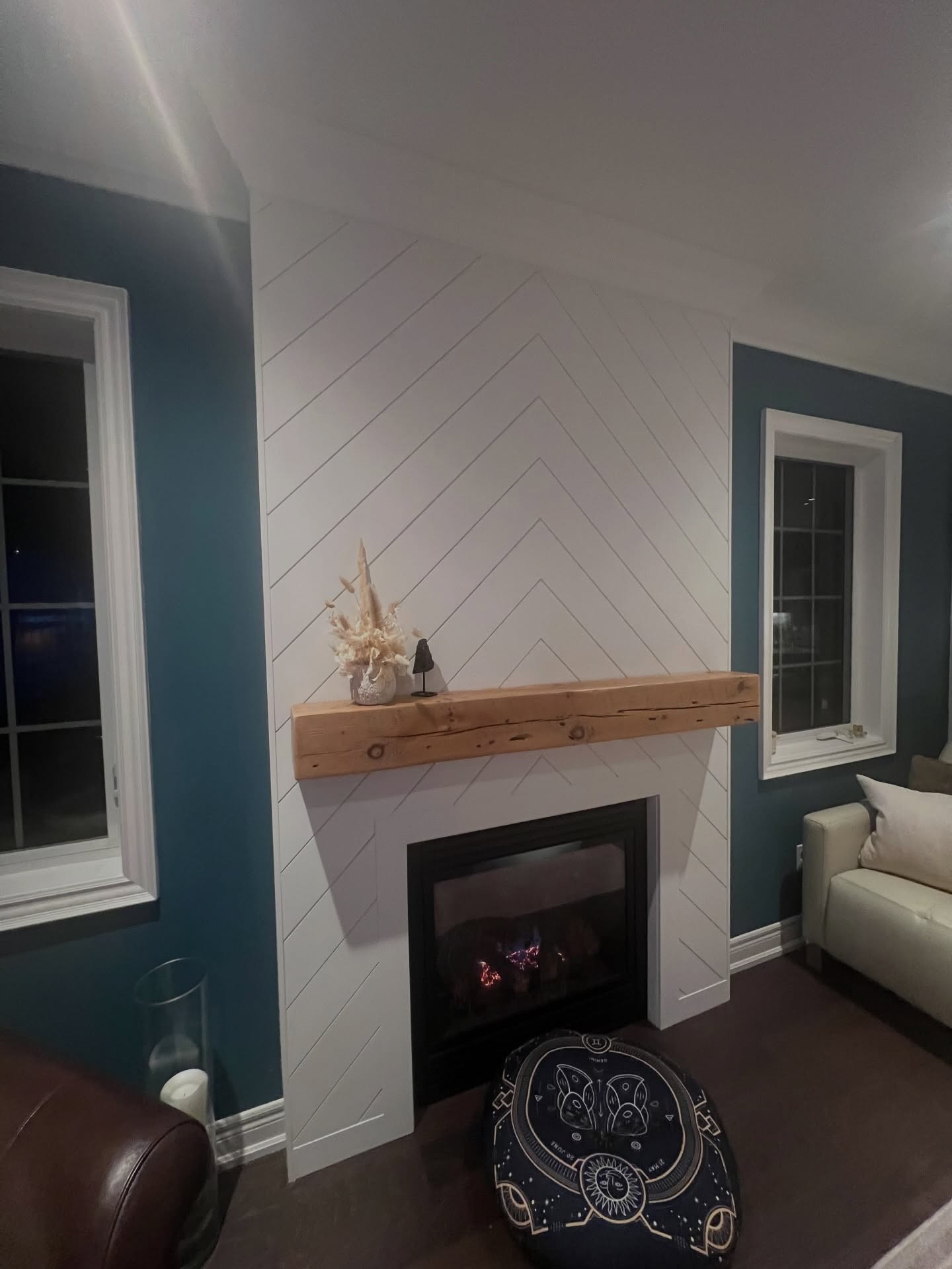 Angled view of the finished chevron fireplace glowing warm at night with the reclaimed barn beam, decor on top, and the dark teal walls now framing the white instead of swallowing it.