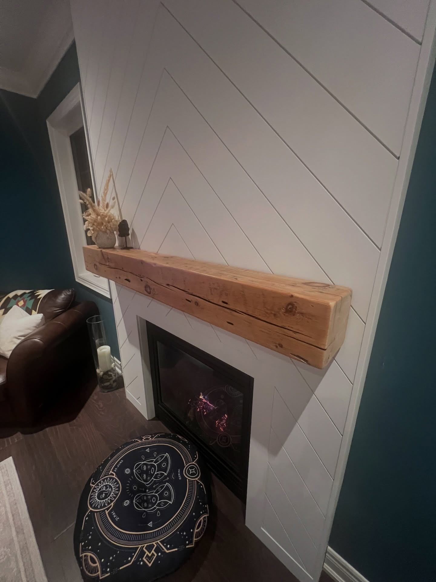 Close-up angled view of the chunky reclaimed barn beam resting on the chevron wall with visible knots, saw marks, and 200 years of character, with the lit gas fire below and a decorative pouf on the floor.