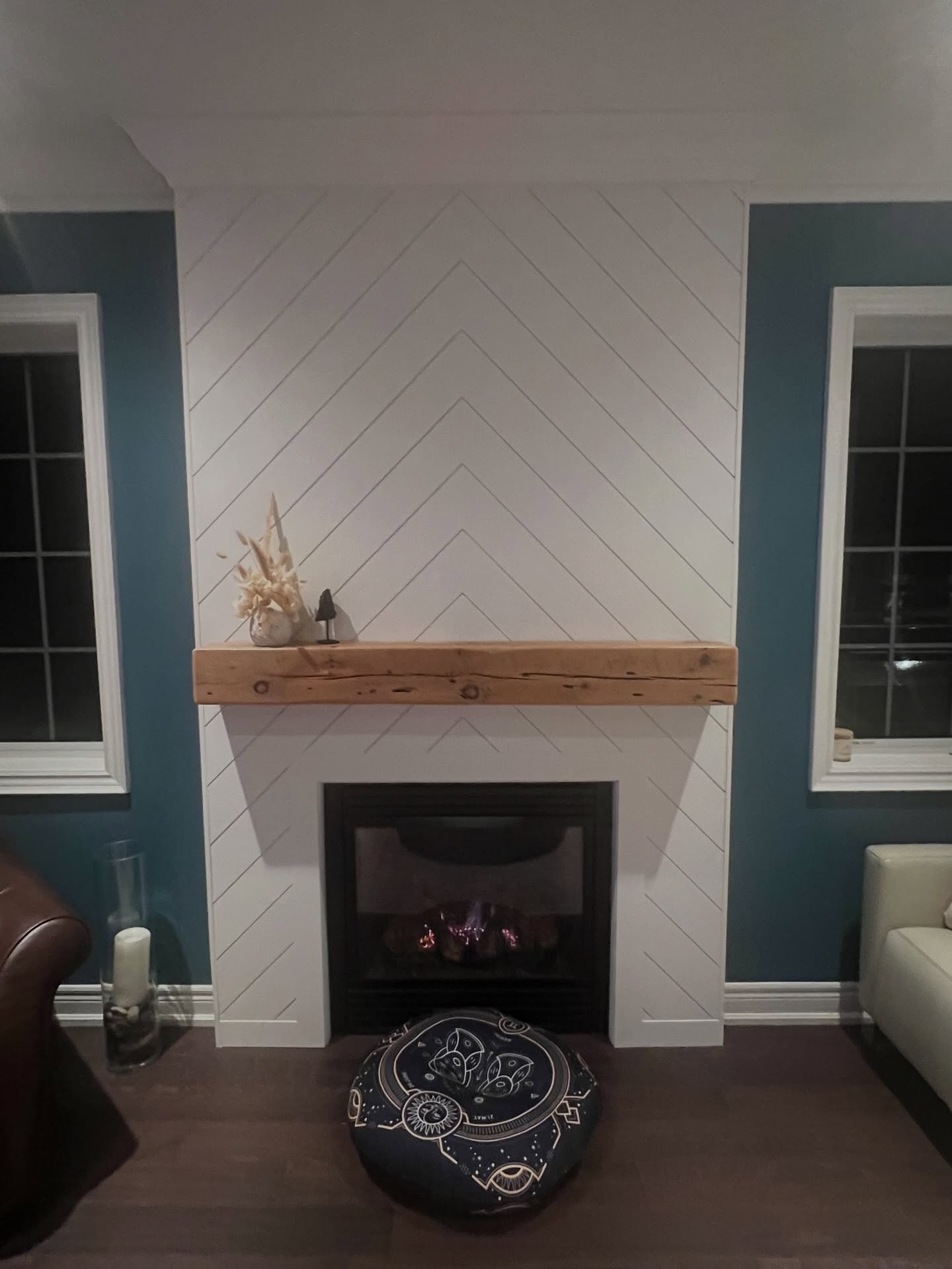 Finished floor-to-ceiling white chevron fireplace with a chunky reclaimed barn beam mantel topped with dried pampas grass and a small sculpture, a lit gas fire glowing in the black firebox, and a round zodiac pouf on the dark wood floor.
