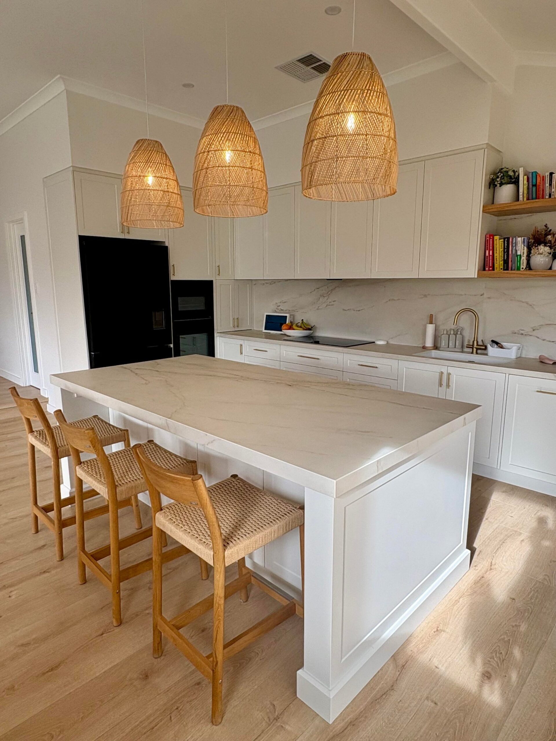Close view of the kitchen island in cream Dekton with soft veining, three rattan pendants above, white Shaker cabinets, and brass handles.