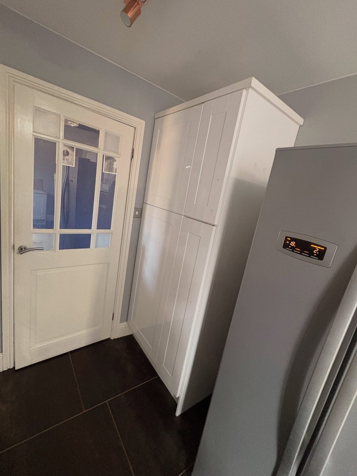 Plain white freestanding larder cabinet beside a tall stainless American fridge-freezer and a white glazed door, over a dark tile floor.