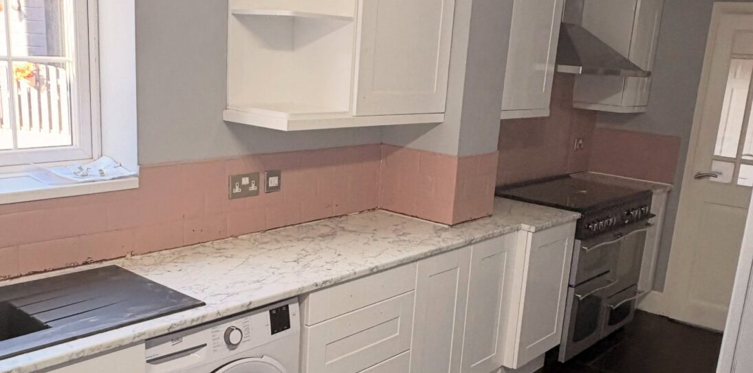 Wide landscape view of a white gloss shaker kitchen before a Stiffkey Blue respray, showing open corner wall cabinets, a stainless hood over a black range cooker, pink subway tile backsplash band, white marble-look counter, built-in Beko washer, and dark grey floor tiles.