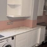 Wide landscape view of a white gloss shaker kitchen before a Stiffkey Blue respray, showing open corner wall cabinets, a stainless hood over a black range cooker, pink subway tile backsplash band, white marble-look counter, built-in Beko washer, and dark grey floor tiles.