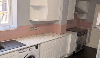 Wide landscape view of a white gloss shaker kitchen before a Stiffkey Blue respray, showing open corner wall cabinets, a stainless hood over a black range cooker, pink subway tile backsplash band, white marble-look counter, built-in Beko washer, and dark grey floor tiles.