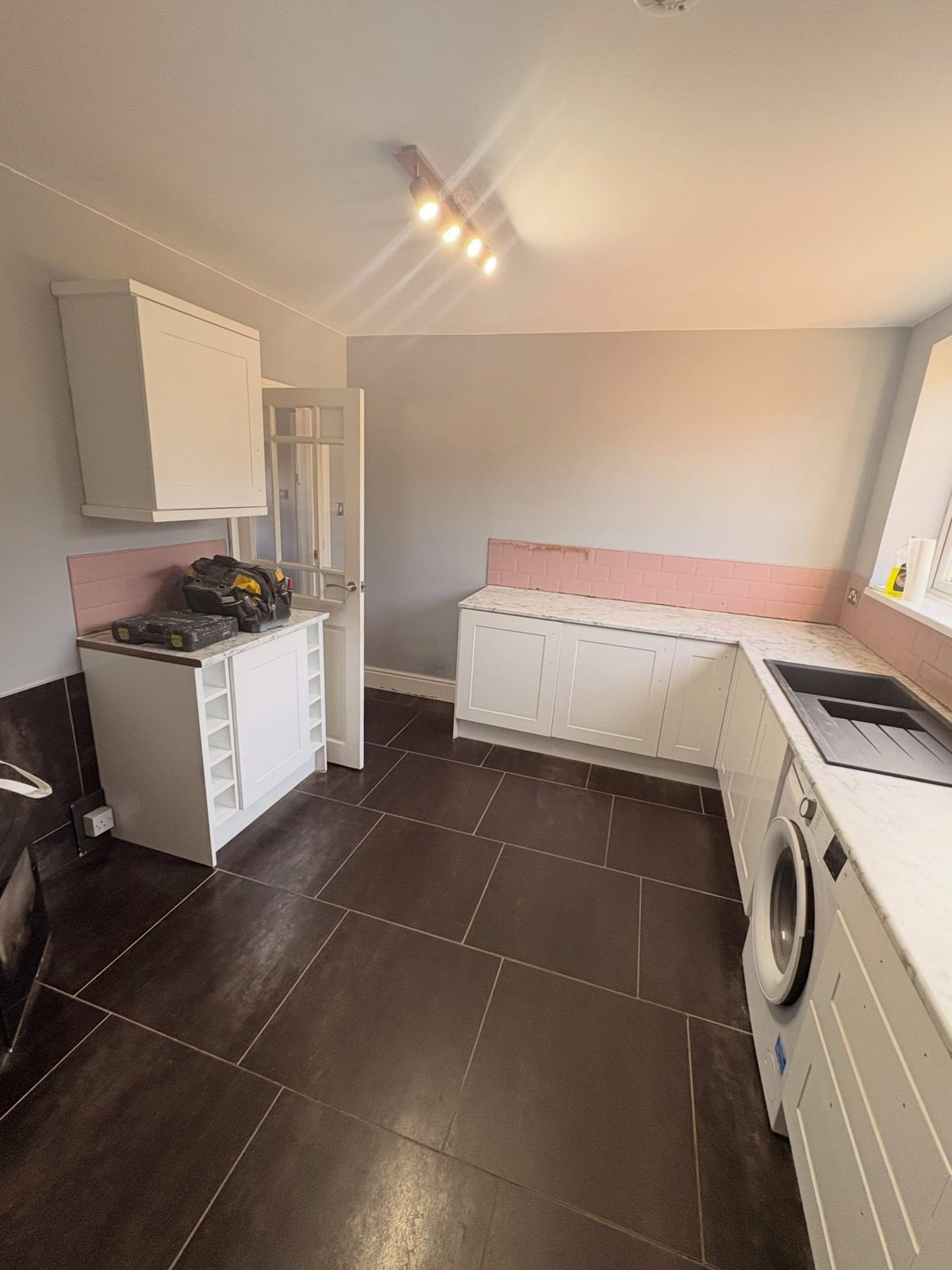 White gloss shaker cabinets with chrome handles on an L-shape run, pink subway tile backsplash, white marble-look counter with a black sink and washer built in, over a dark grey tile floor.