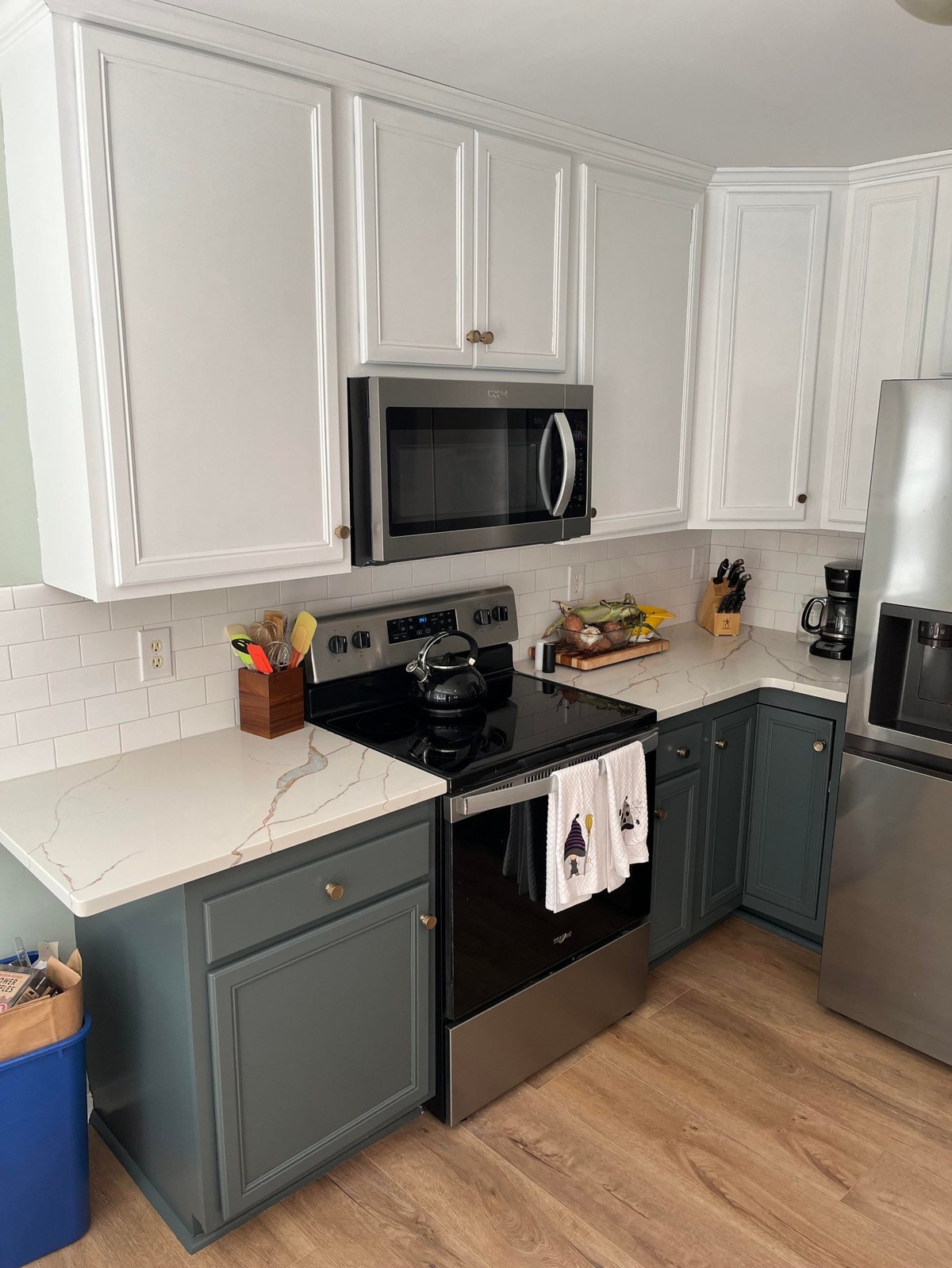 After photo of the stove corner with white upper cabinets, dark gray-green Flint lower cabinets, white subway tile backsplash, Calcutta Oro Leaf white quartz counters with soft gold veining, a stainless Whirlpool over-the-range microwave, and oak LVP flooring.