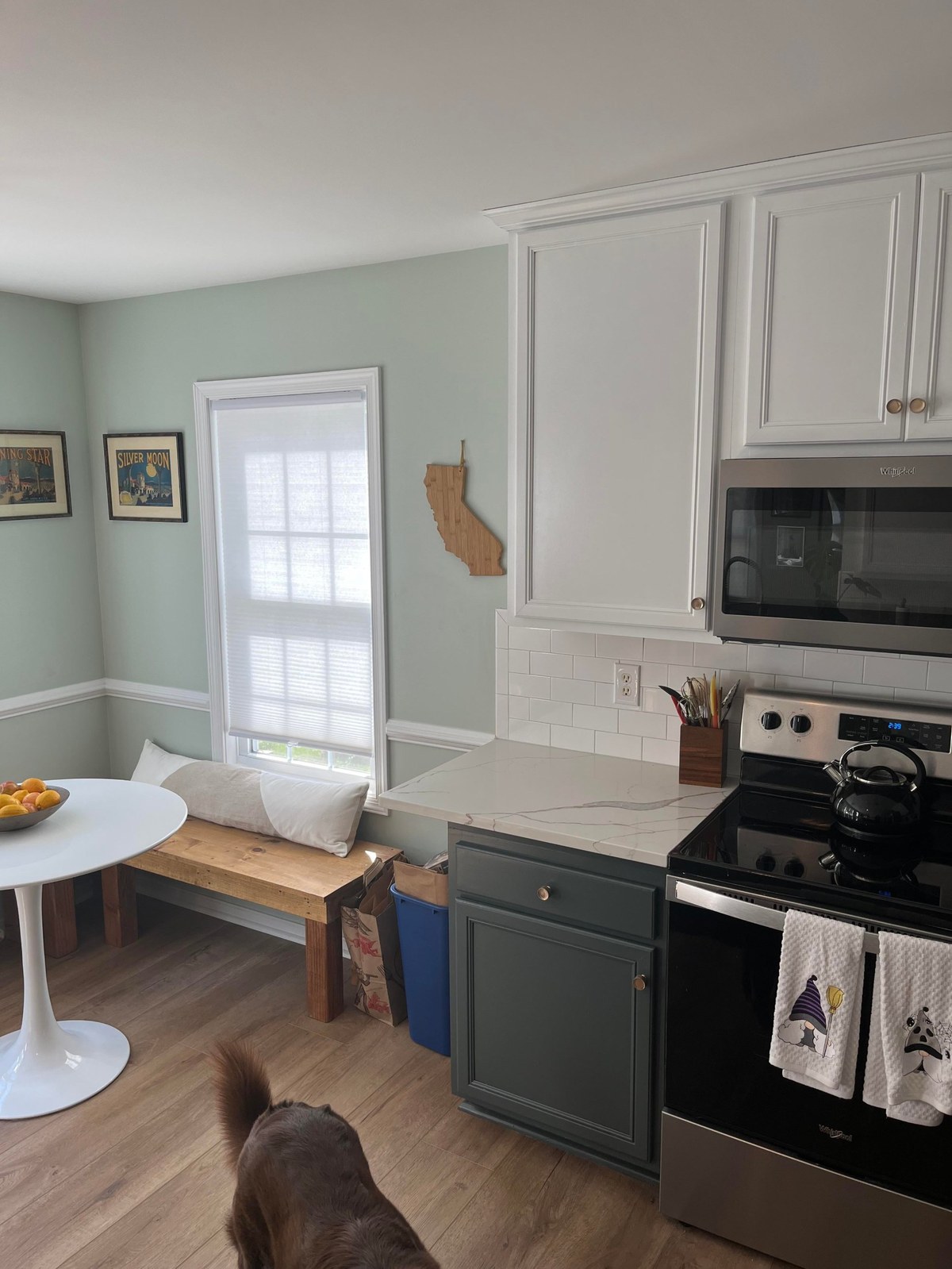 After photo of the breakfast nook with a raw softwood L-shaped bench against the sage green wall, a white Tulip-style table with a bowl of fresh fruit, two framed vintage lithograph posters on the wall, a cordless window shade, and the tail of a chocolate dog visible on the oak LVP floor.