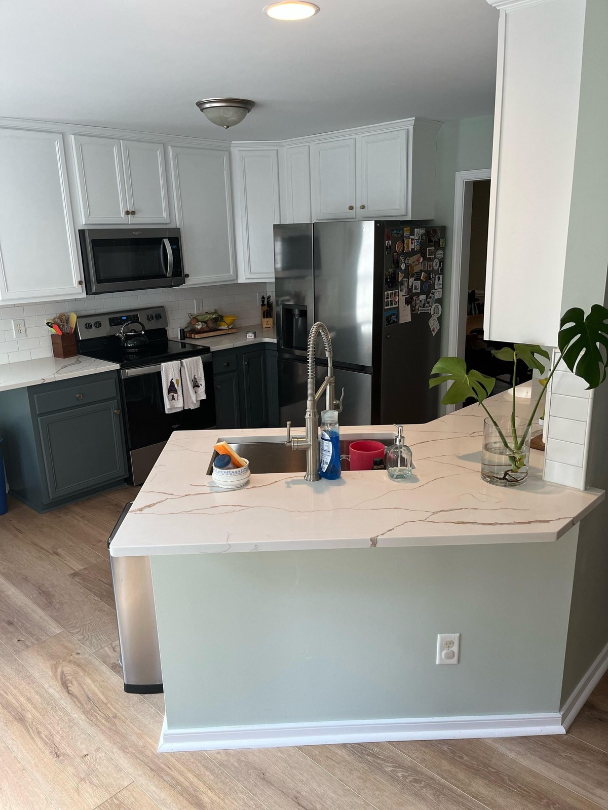 Wide after view of the kitchen with white upper cabinets, Flint gray-green lower cabinets, white subway tile backsplash, Calcutta Oro Leaf quartz counters, oak LVP flooring, a stainless Whirlpool microwave, the original black stainless LG side-by-side fridge, and the sage walls finally reading as a calm backdrop.