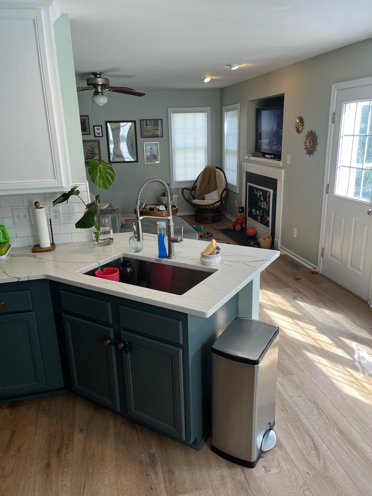 After photo of the kitchen peninsula with dark gray-green painted cabinets, a thick Calcutta Oro Leaf quartz countertop with gold veining, a deep single bowl undermount sink, the original tall spring-arm pull-down faucet, and a view into the sage green living room beyond with the ceiling fan and gallery wall still in place.
