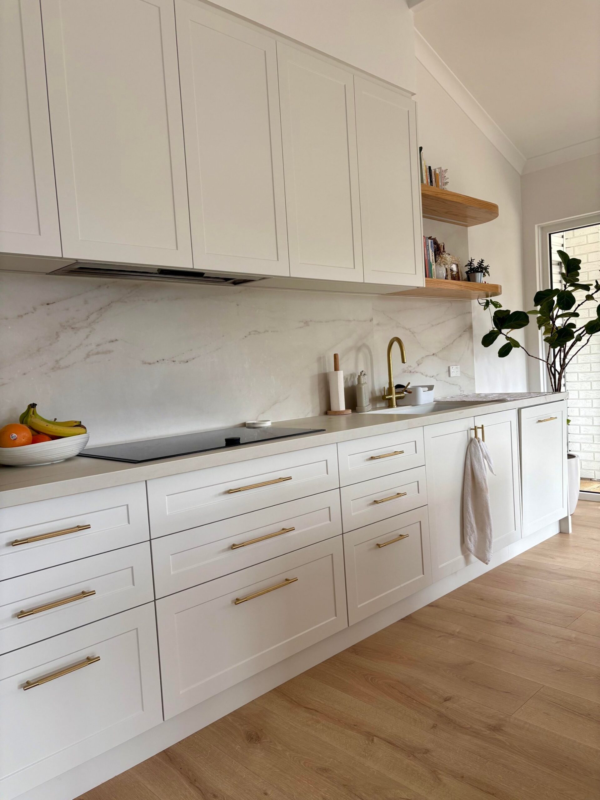 Cooktop wall with white Shaker cabinets, brass pulls, a brass goose-neck tap, open timber shelves with cookbooks, and a marble-look backsplash.