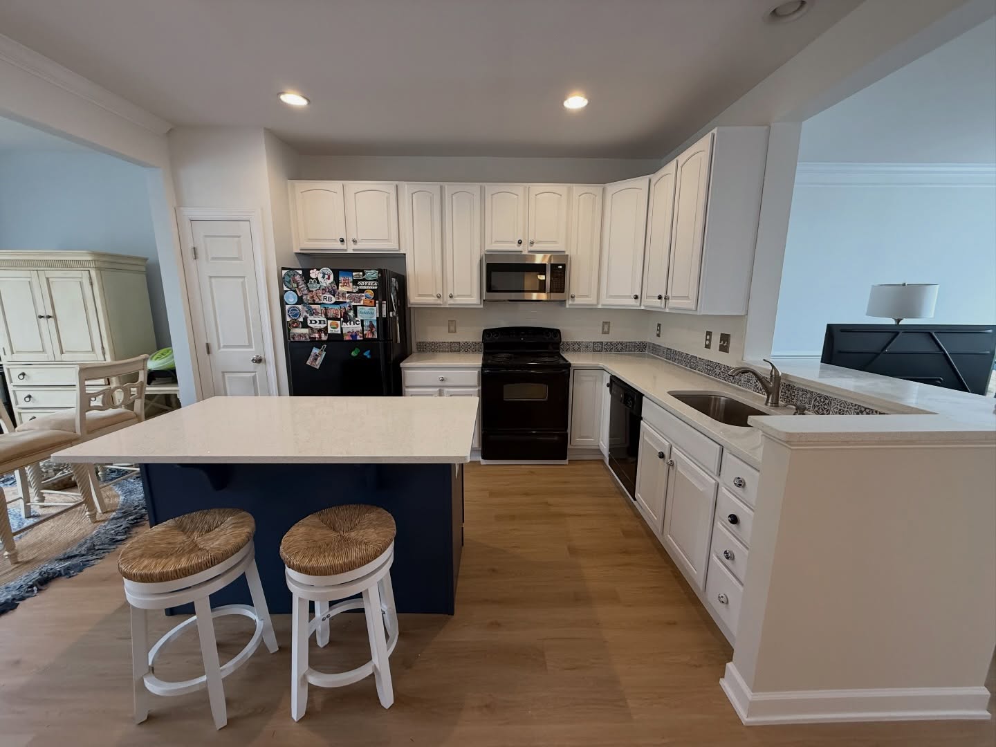 Wide view of the kitchen from the dining side, showing the navy Indigo Batik island with two round rush-seat stools, white perimeter cabinets, a black fridge, a black stove, a stainless microwave, a white pantry door, and light oak flooring.