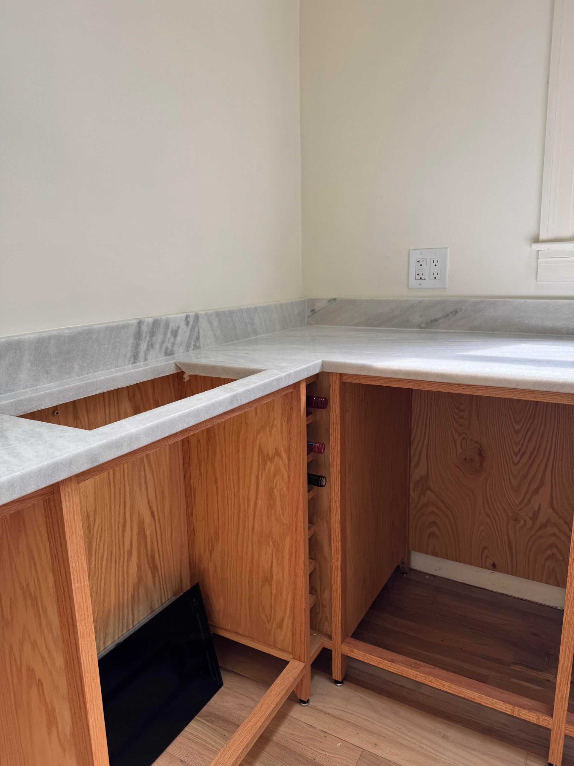 White marble L-shaped counter with gray veining installed on raw oak cabinets, with a small vertical wine bottle rack built between two bays.