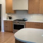 White oak kitchen with a curved fluted island, white quartz countertop, stainless gas range, plaster hood, and tall built-in fridge in the background.