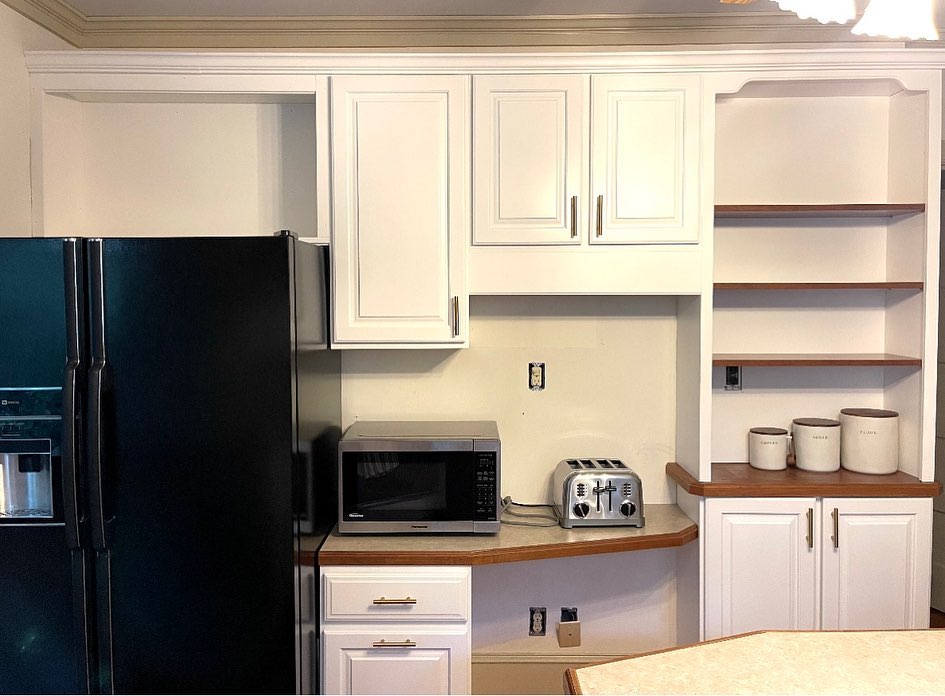 Same kitchen corner after the makeover, with the upper cabinets and bookshelf column painted satin white, the open bookshelf shelves re-stained a darker walnut tone against the white, a stainless Panasonic microwave in the nook, three ceramic canisters labeled coffee, sugar, and flour on the counter, and the same black side-by-side fridge still in place.