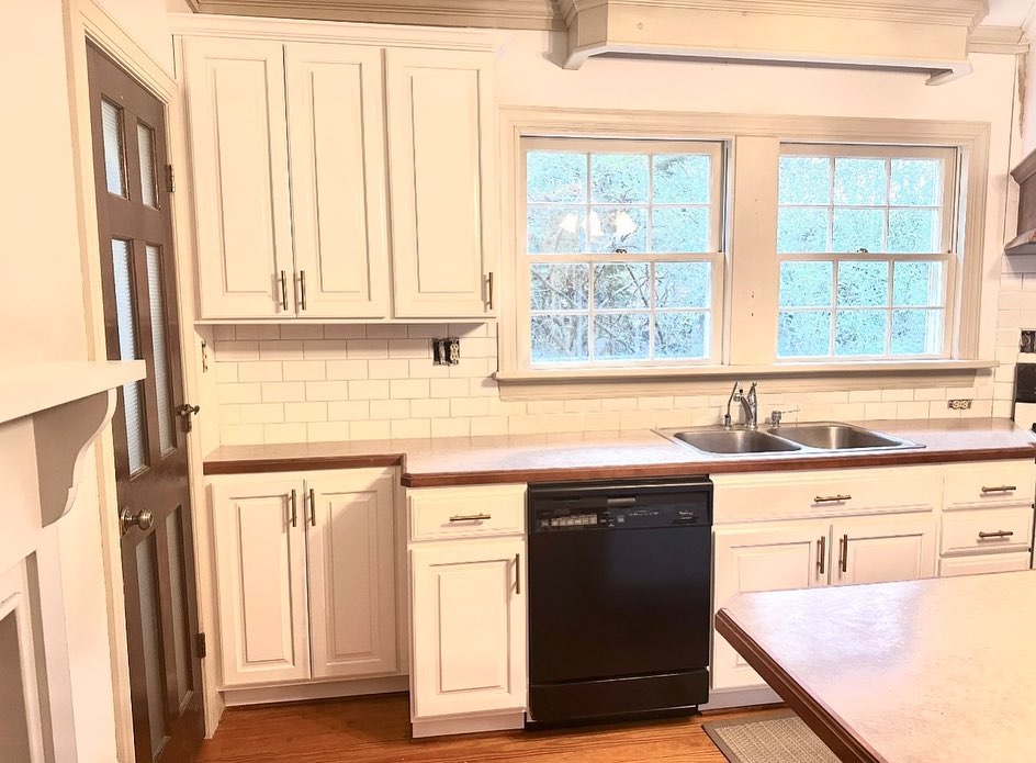 Same kitchen sink wall after the makeover, with cabinets painted satin white, a white 3x6 inch subway tile backsplash, brushed nickel bar pulls, a dark wood edge band on the formica counter, and the same black Whirlpool dishwasher kept in place.