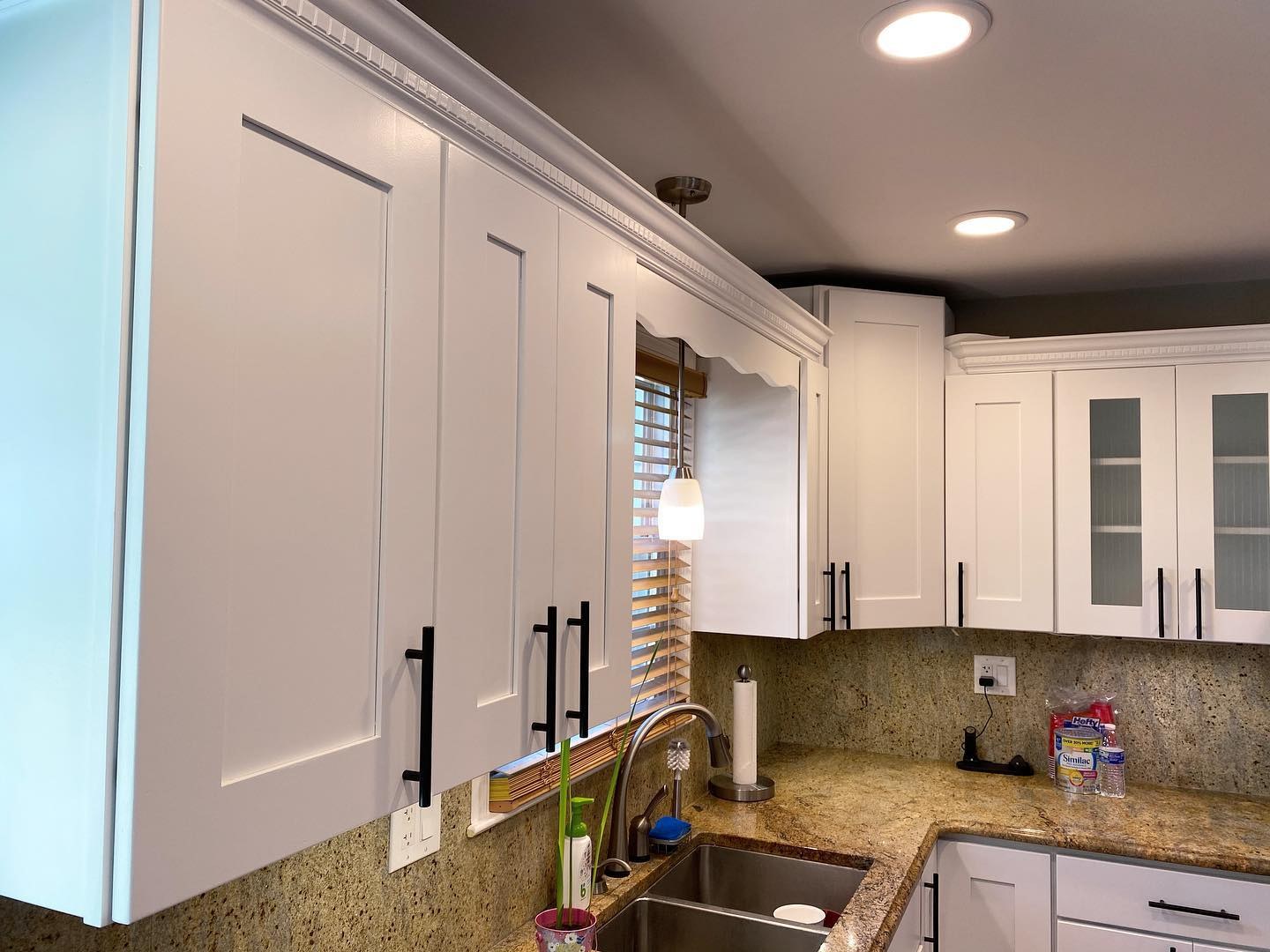 Close up of white painted shaker cabinets with dentil crown molding, matte black bar pulls, a small pendant light, and the original speckled granite counter and backsplash.