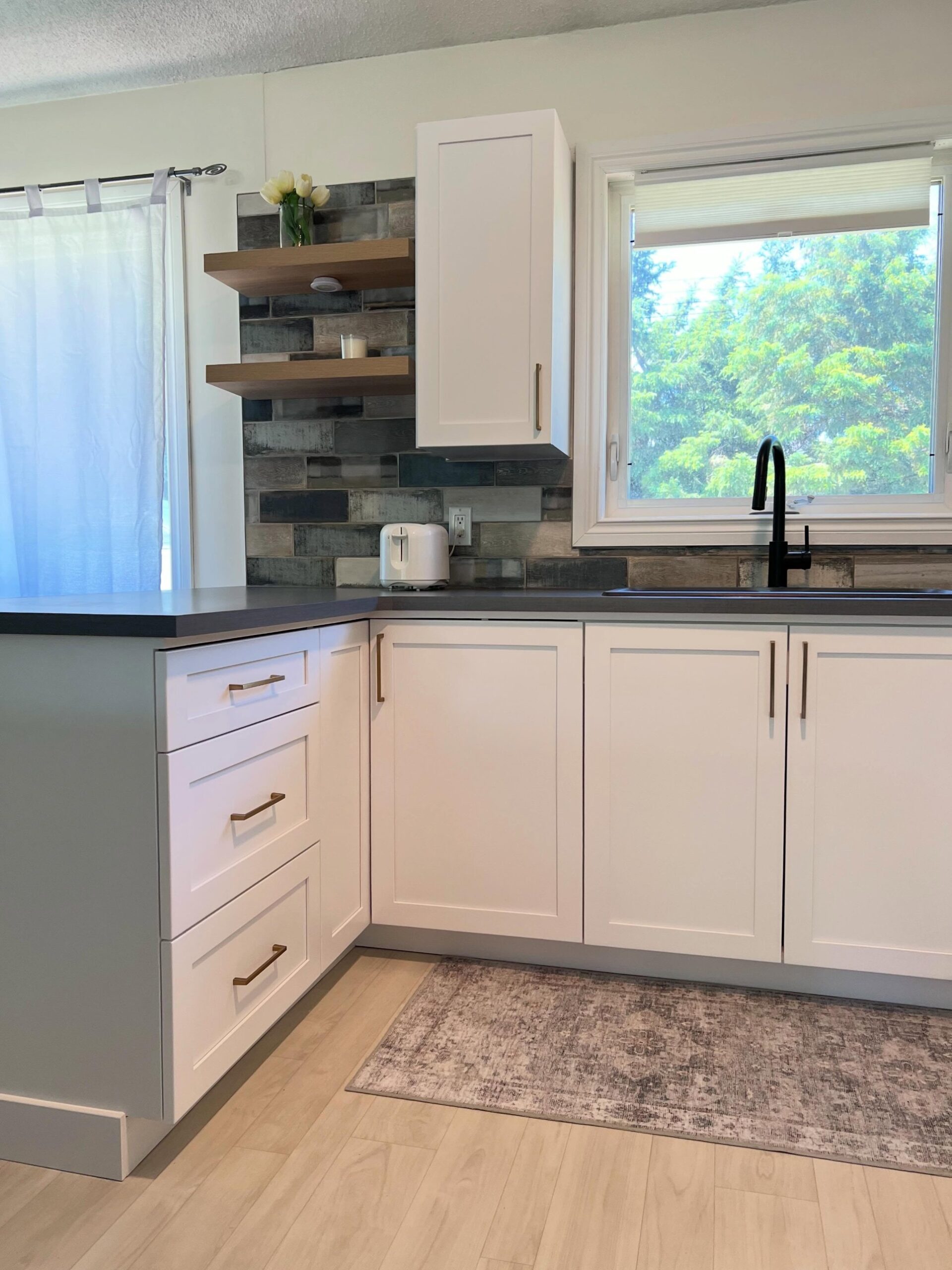 Side view showing Chantilly Lace Shaker drawers with gold bar pulls, the dark gray counter, floating oak shelves styled with white tulips and a candle, and a soft layered rug on blonde flooring.
