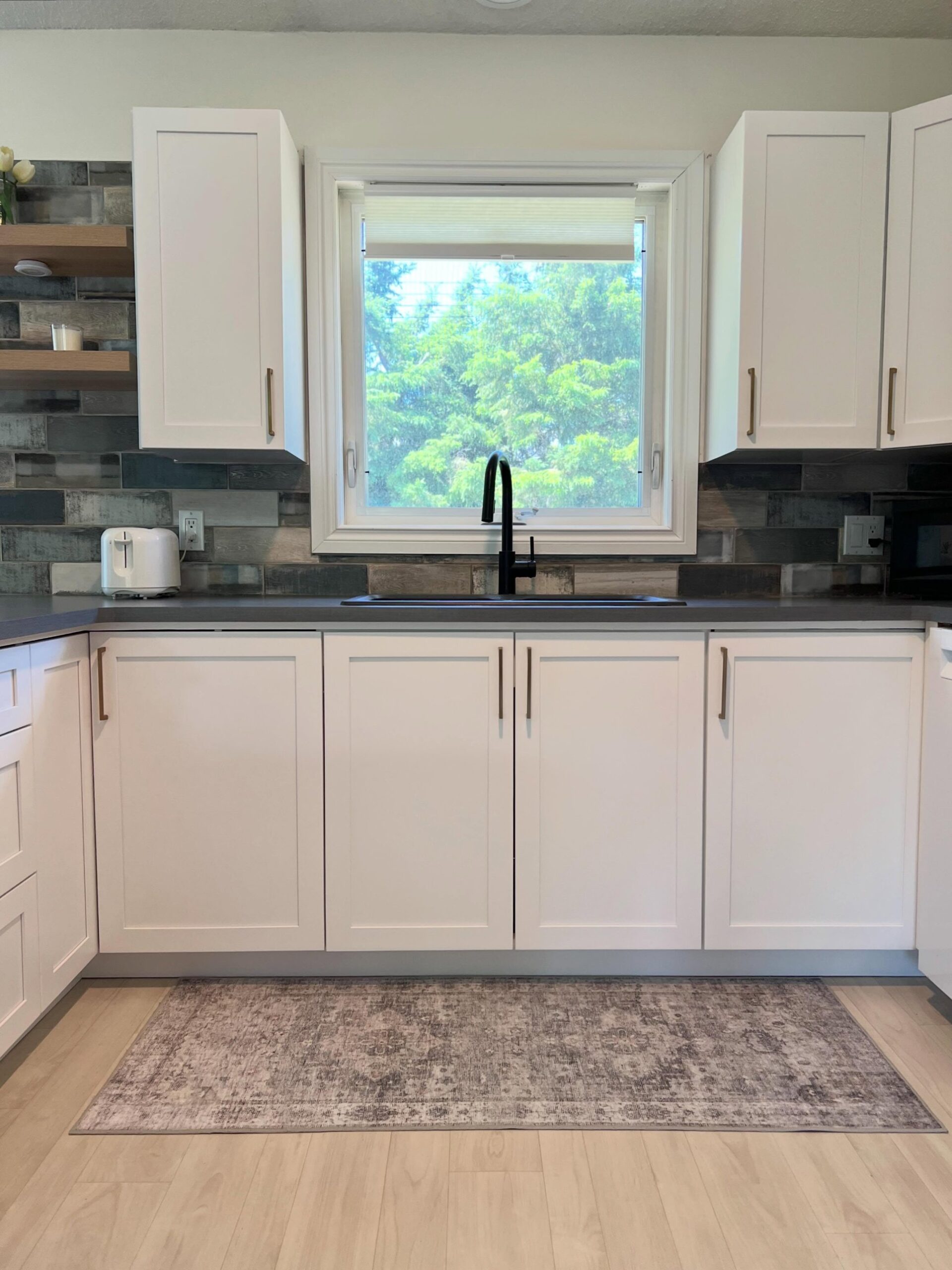 Head on view of the sink wall after refacing with Chantilly Lace Shaker cabinets, gold bar pulls, a black gooseneck faucet, a single-bowl black sink, and a vintage style area rug on blonde LVP flooring.