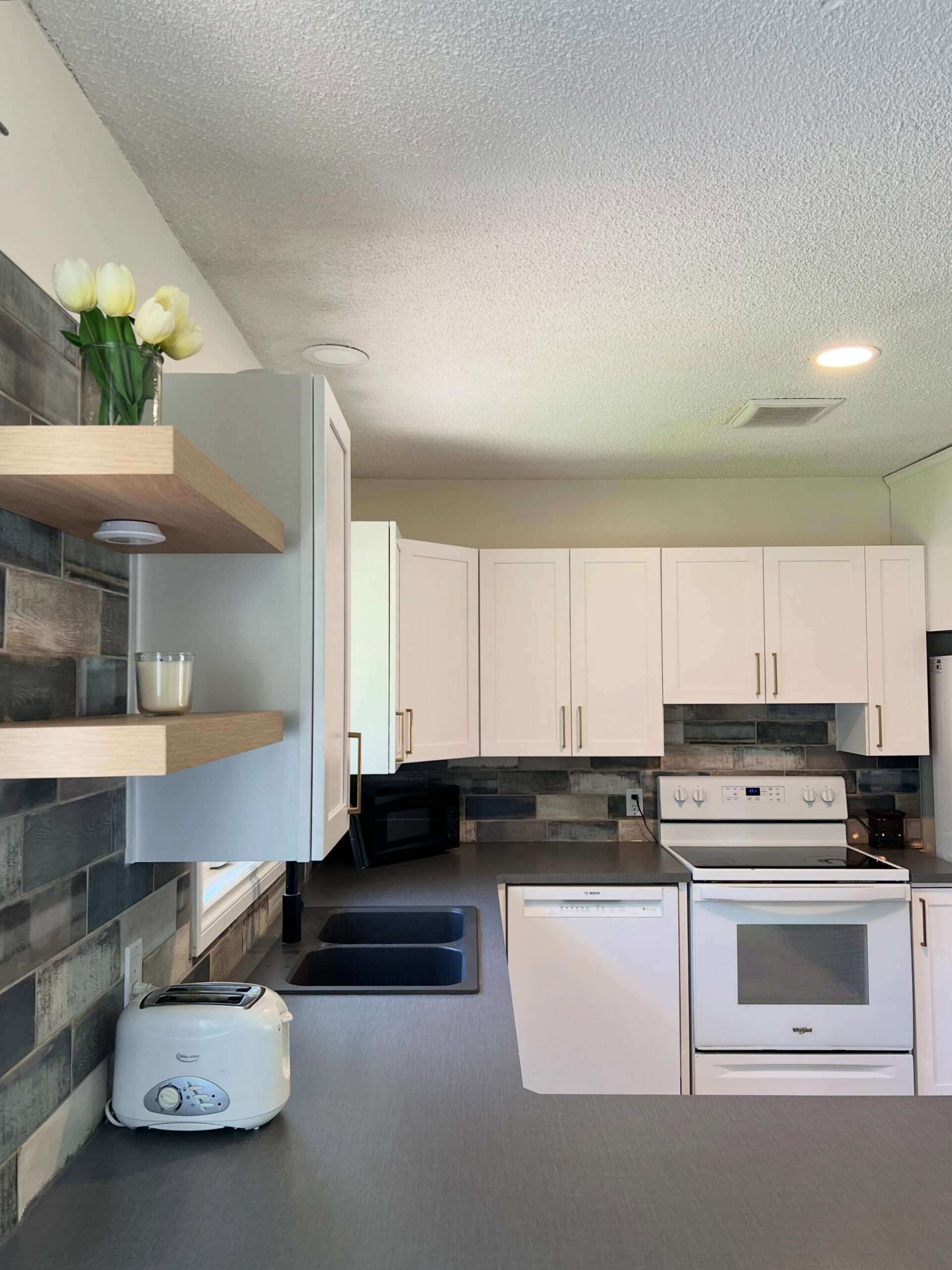 Interior angle of the finished kitchen showing floating oak shelves styled with tulips and a candle, Chantilly Lace Shaker cabinets, gold bar pulls, a black double sink, white appliances, and the rustic tile running wall to wall.
