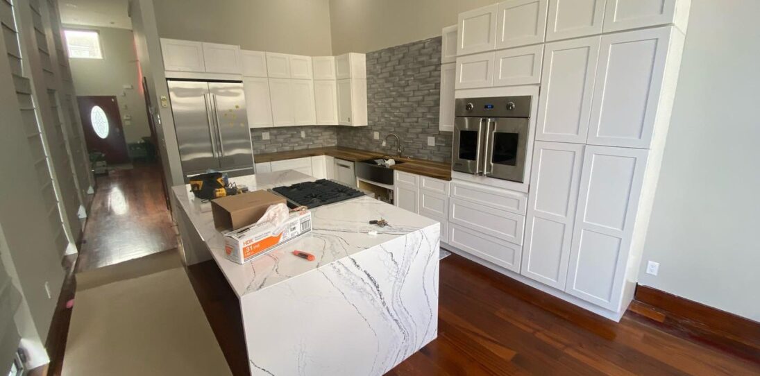 Wide view of a brand-new white shaker kitchen before painting, with a Cambria quartz waterfall island in the center, tall white cabinets flanking a stainless Viking wall oven, grey glazed brick backsplash, built-in stainless fridge, and dark cherry wood floors throughout.