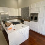 Wide view of a brand-new white shaker kitchen before painting, with a Cambria quartz waterfall island in the center, tall white cabinets flanking a stainless Viking wall oven, grey glazed brick backsplash, built-in stainless fridge, and dark cherry wood floors throughout.