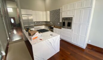 Wide view of a brand-new white shaker kitchen before painting, with a Cambria quartz waterfall island in the center, tall white cabinets flanking a stainless Viking wall oven, grey glazed brick backsplash, built-in stainless fridge, and dark cherry wood floors throughout.