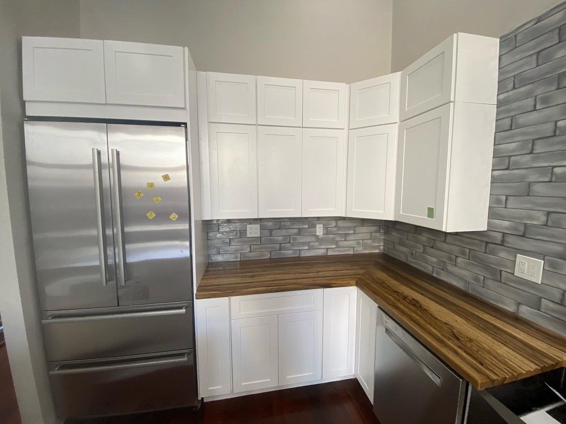 Corner view of the white kitchen with a built-in stainless fridge, white upper cabinets, grey glazed brick backsplash, and warm butcher block counters.