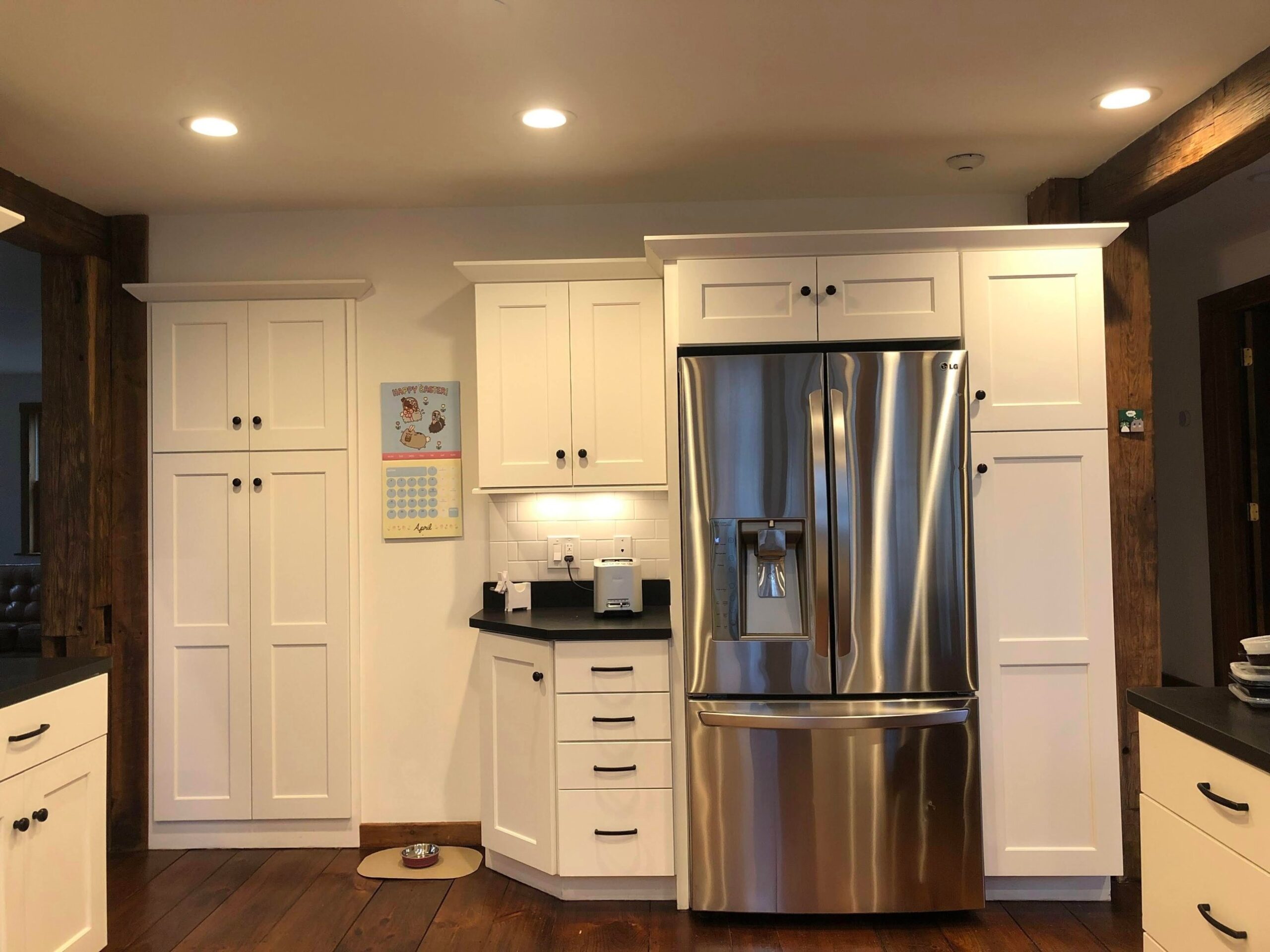 White cabinets and stainless French-door fridge against a white wall, with a wall calendar and a dog bowl on the pine floor.