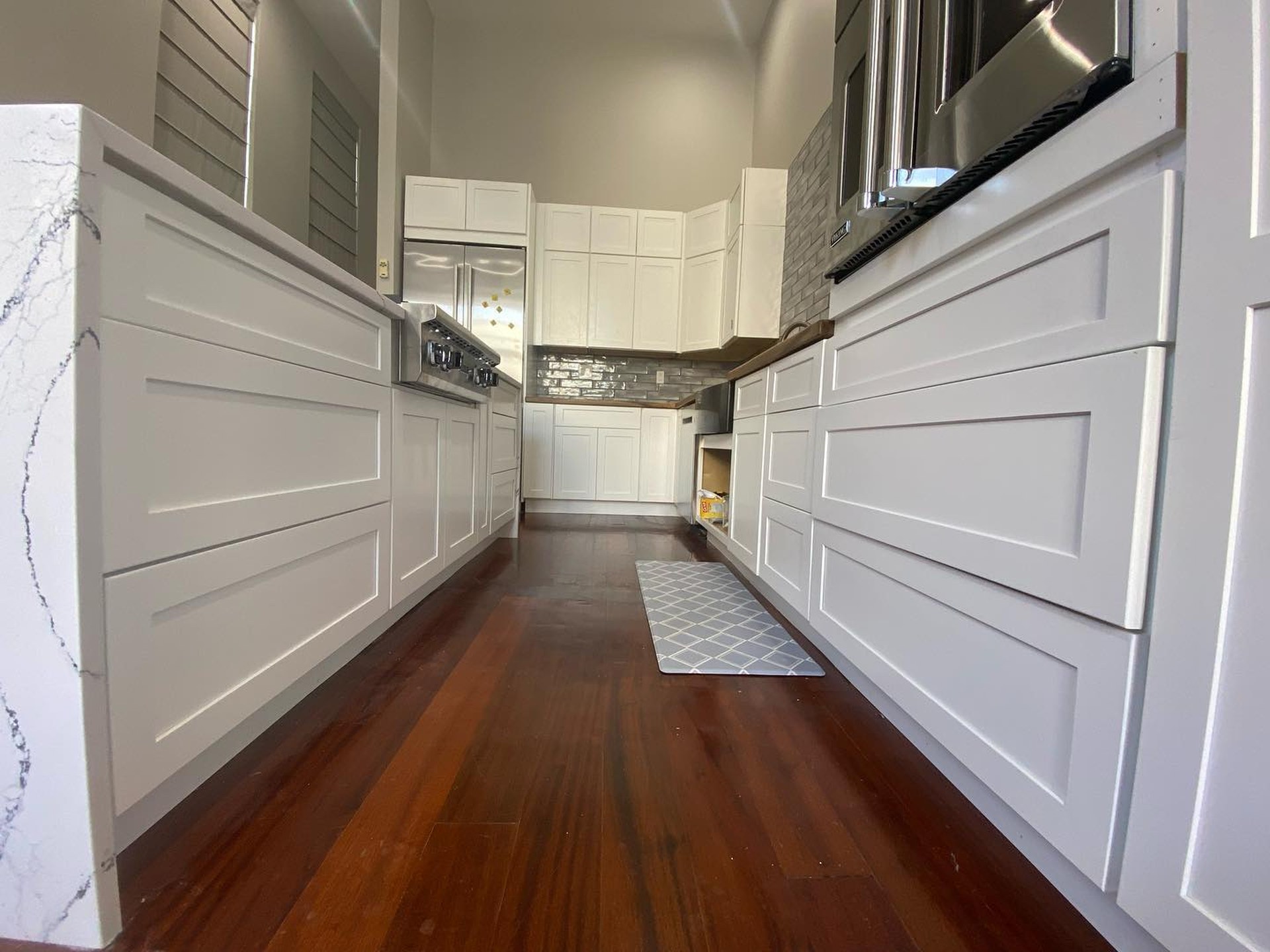 Low angle view down a galley kitchen with white shaker drawer fronts on both sides, Cambria quartz waterfall island on the left, and dark cherry floors leading to a corner cabinet run.