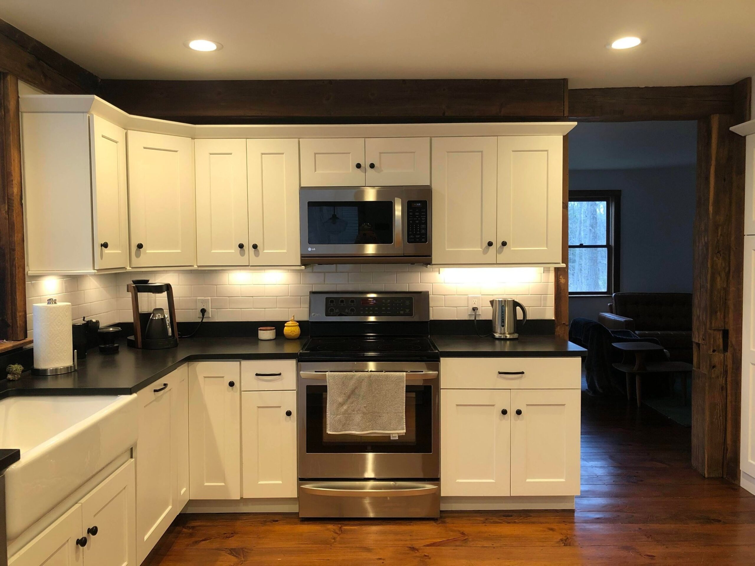 White Shaker uppers over a black counter, white subway tile, a stainless stove and a stainless over-the-range microwave, with heavy wood beams framing the view.