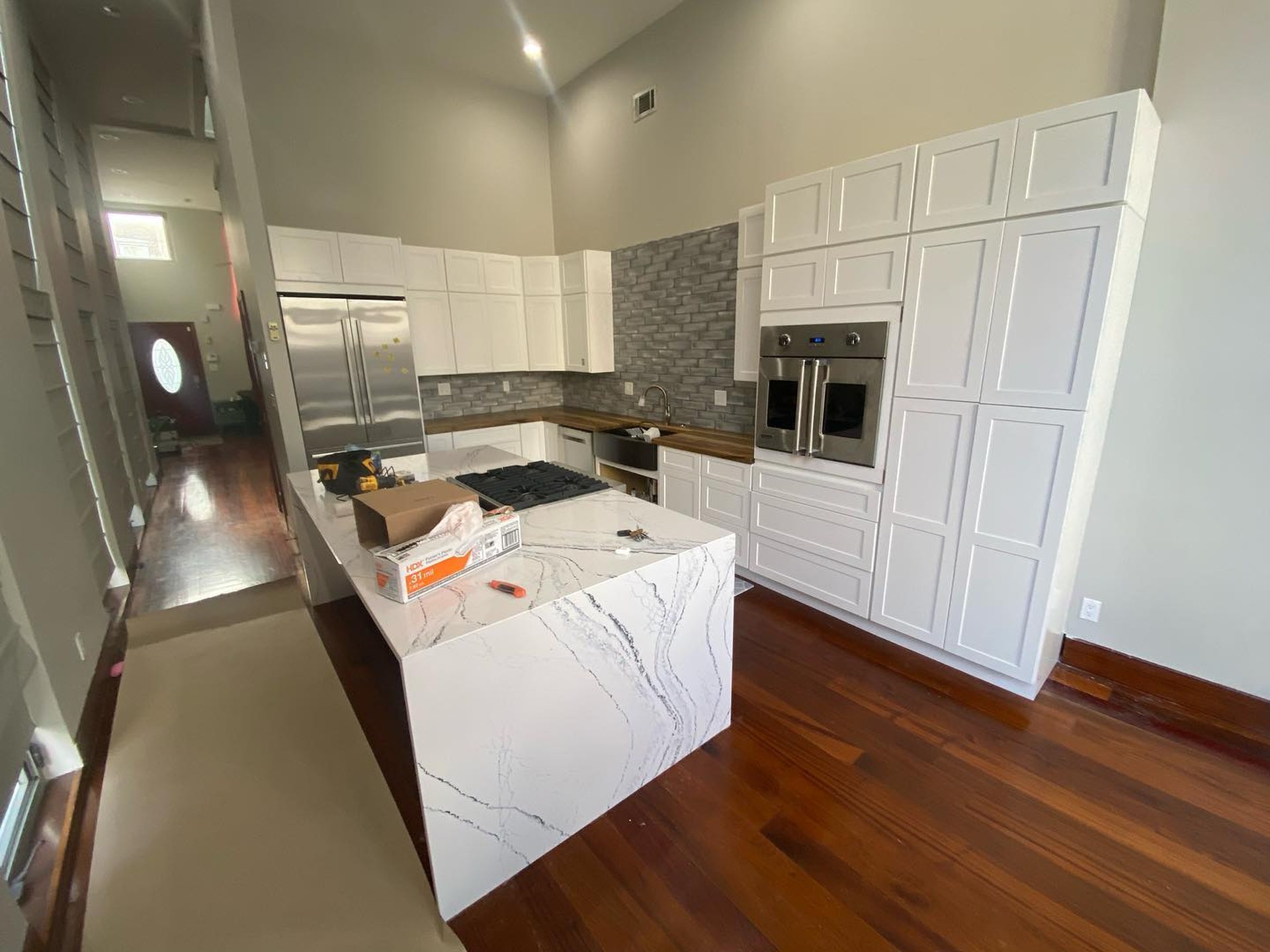Wide view of a tall-ceiling kitchen with brand-new white shaker cabinets, a waterfall-edge Cambria quartz island, grey glazed brick backsplash, and rich cherry wood floors.