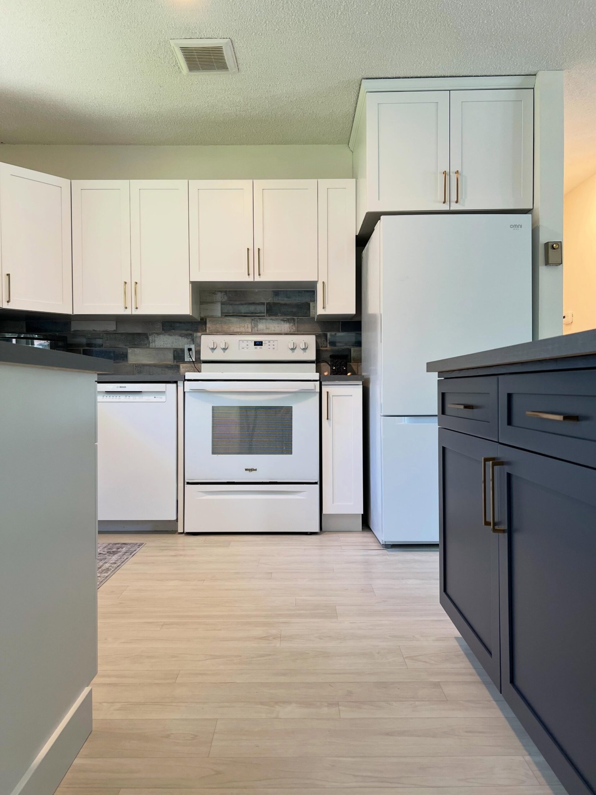 Finished range wall with Chantilly Lace Shaker uppers and gold hardware, a white Whirlpool range, a white Bosch dishwasher, a Hale Navy base cabinet on the right, and blonde LVP flooring running under all of it.