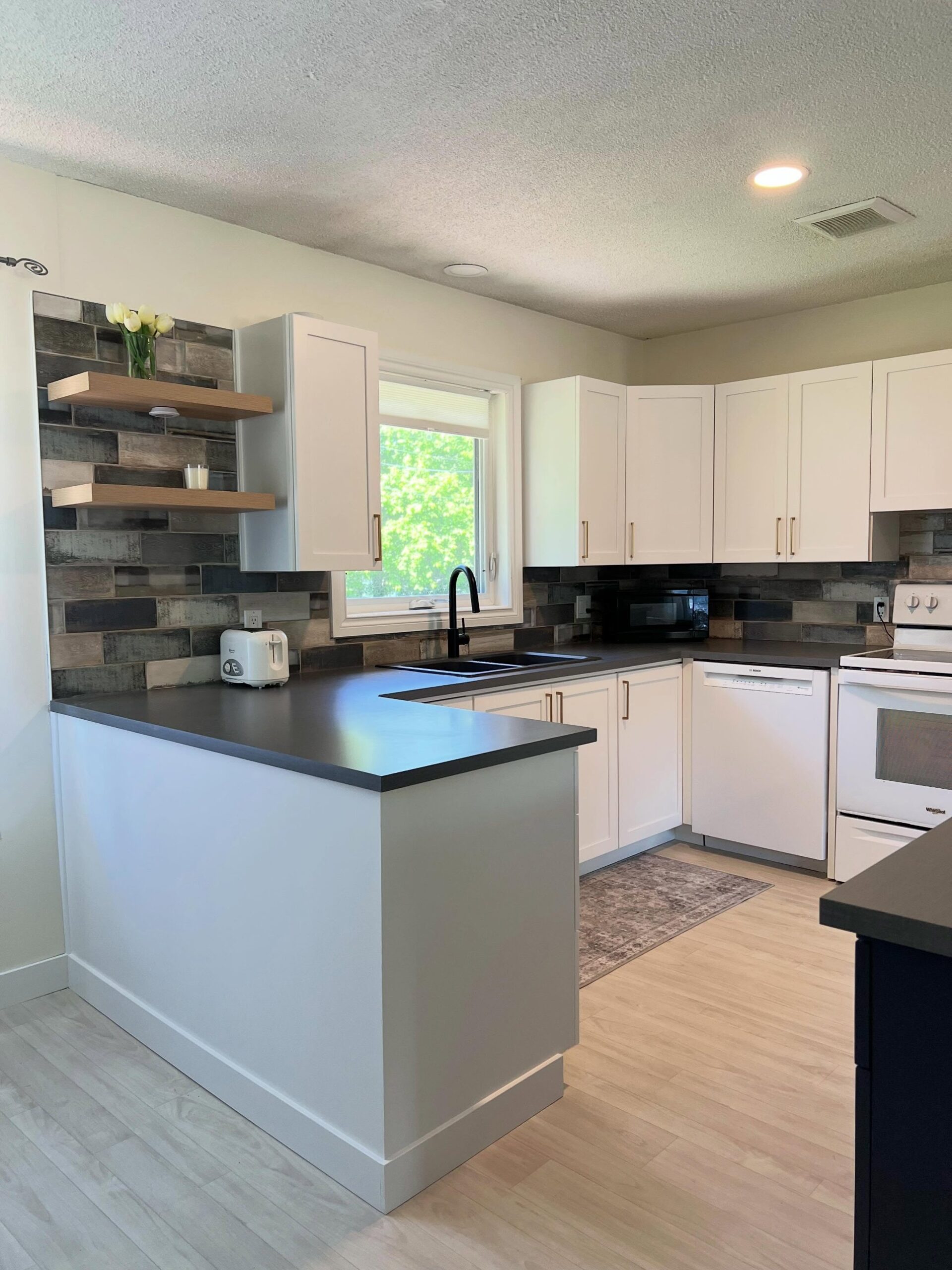 Wide view of the finished U-shaped kitchen with Chantilly Lace Shaker cabinets, a dark gray laminate counter, a navy base cabinet in the foreground, floating oak shelves, the rustic multi-tone tile backsplash, and white Whirlpool and Bosch appliances.