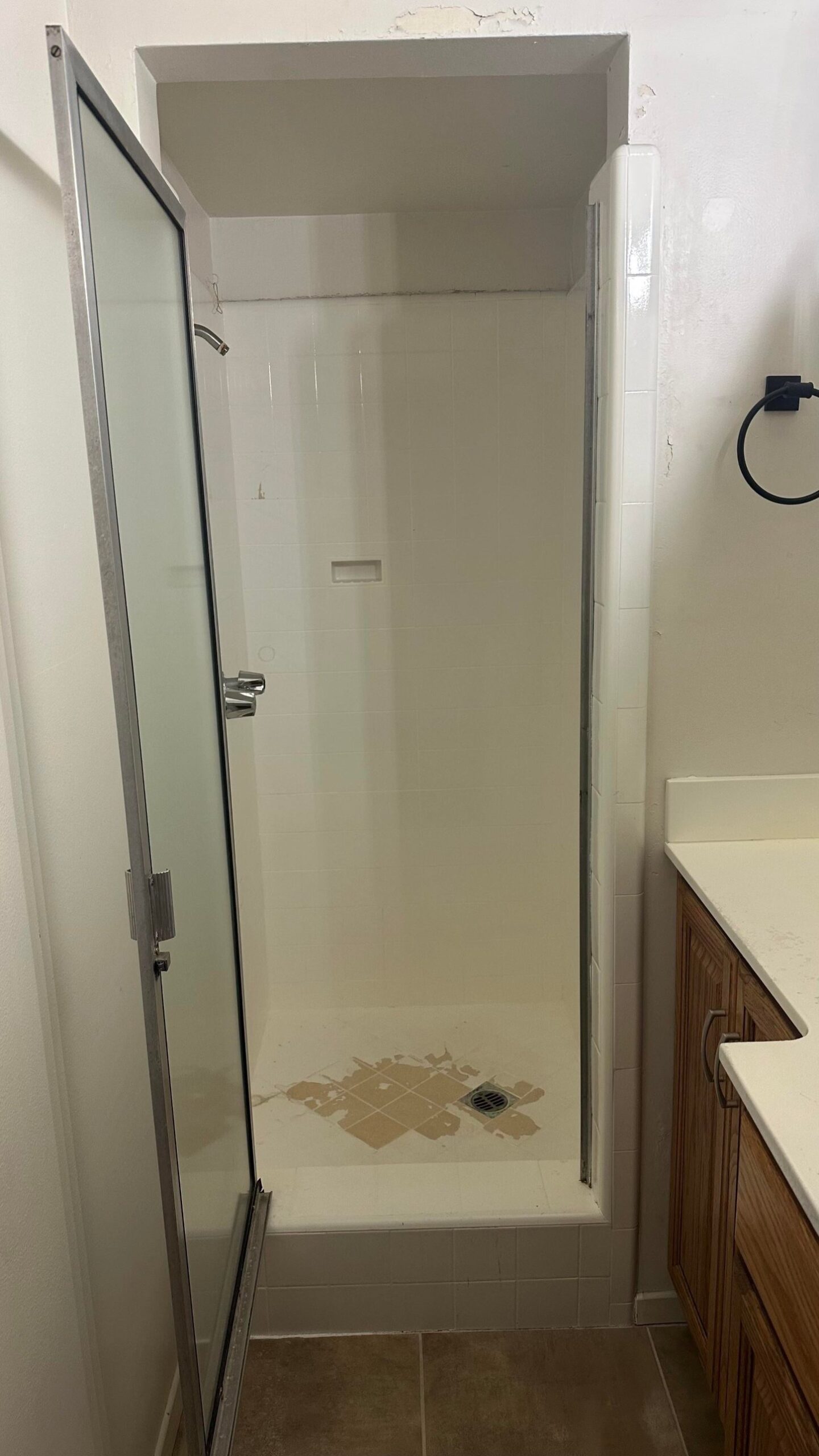 Straight on view into a narrow white tile shower with a chrome valve handle, bare walls, and a damaged tile floor around the drain.