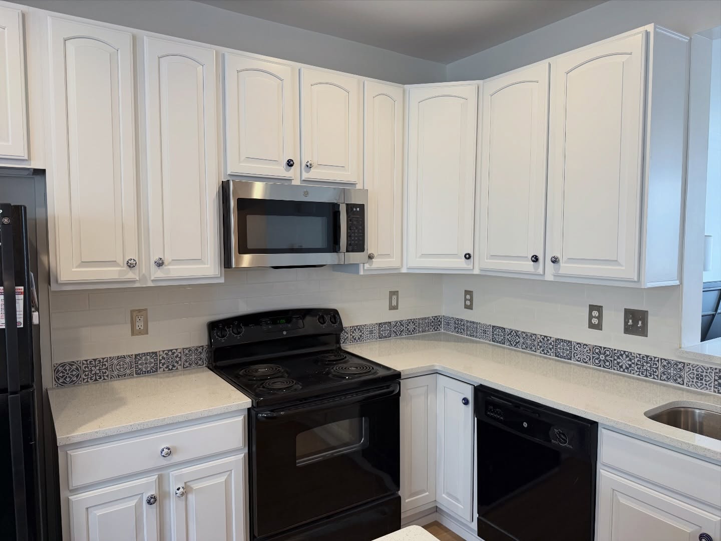 Kitchen with white arched cabinets painted Sherwin-Williams Pure White, a black electric coil range, a stainless microwave, white quartz counters, and the original Moroccan tile strip now reading as a decorative feature.