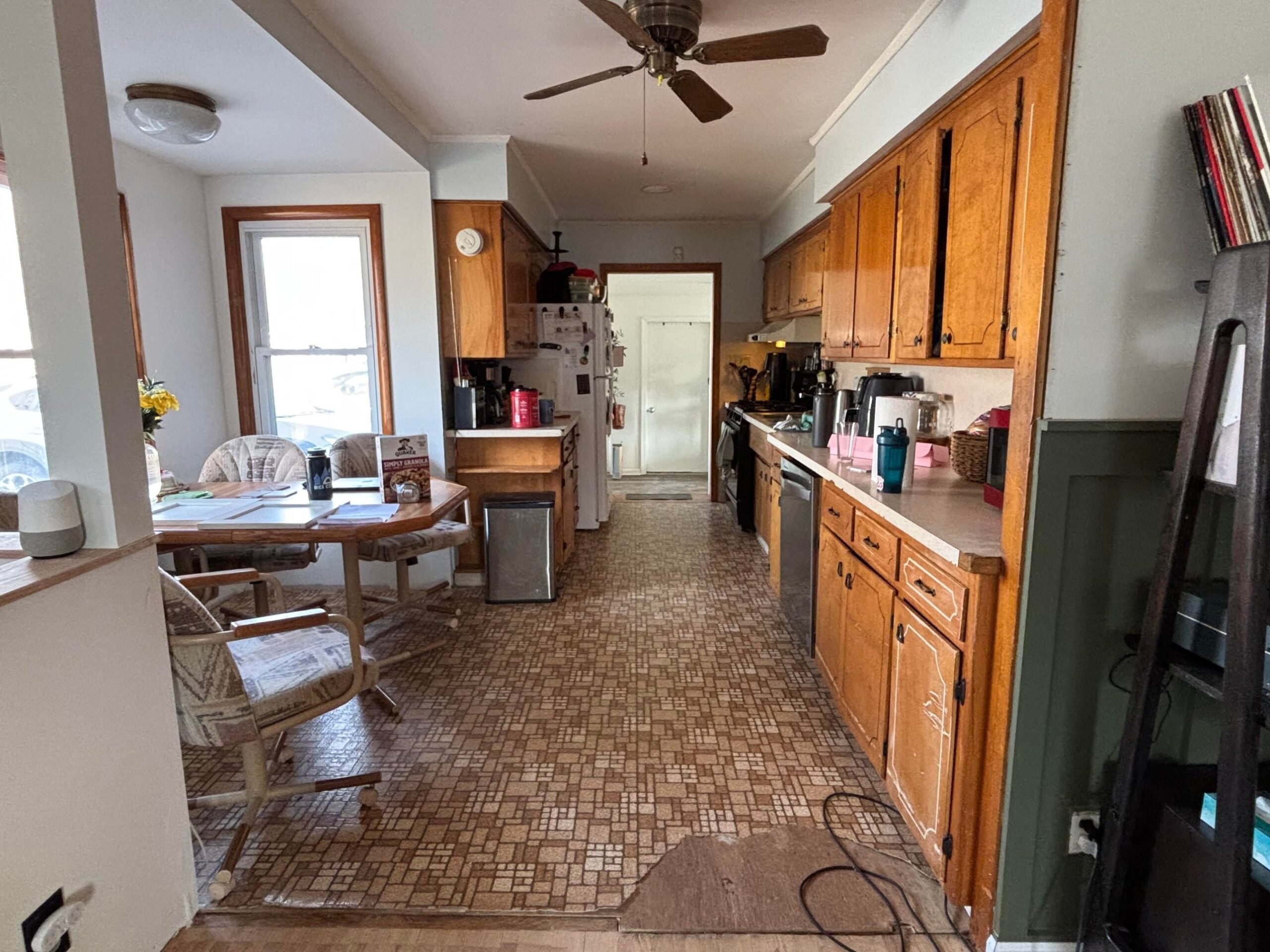 Wide view down a galley kitchen with orange oak cabinets, brown patterned linoleum peeling at the edge, a ceiling fan, and a breakfast nook with swivel chairs and the octagon table visible at the far left.