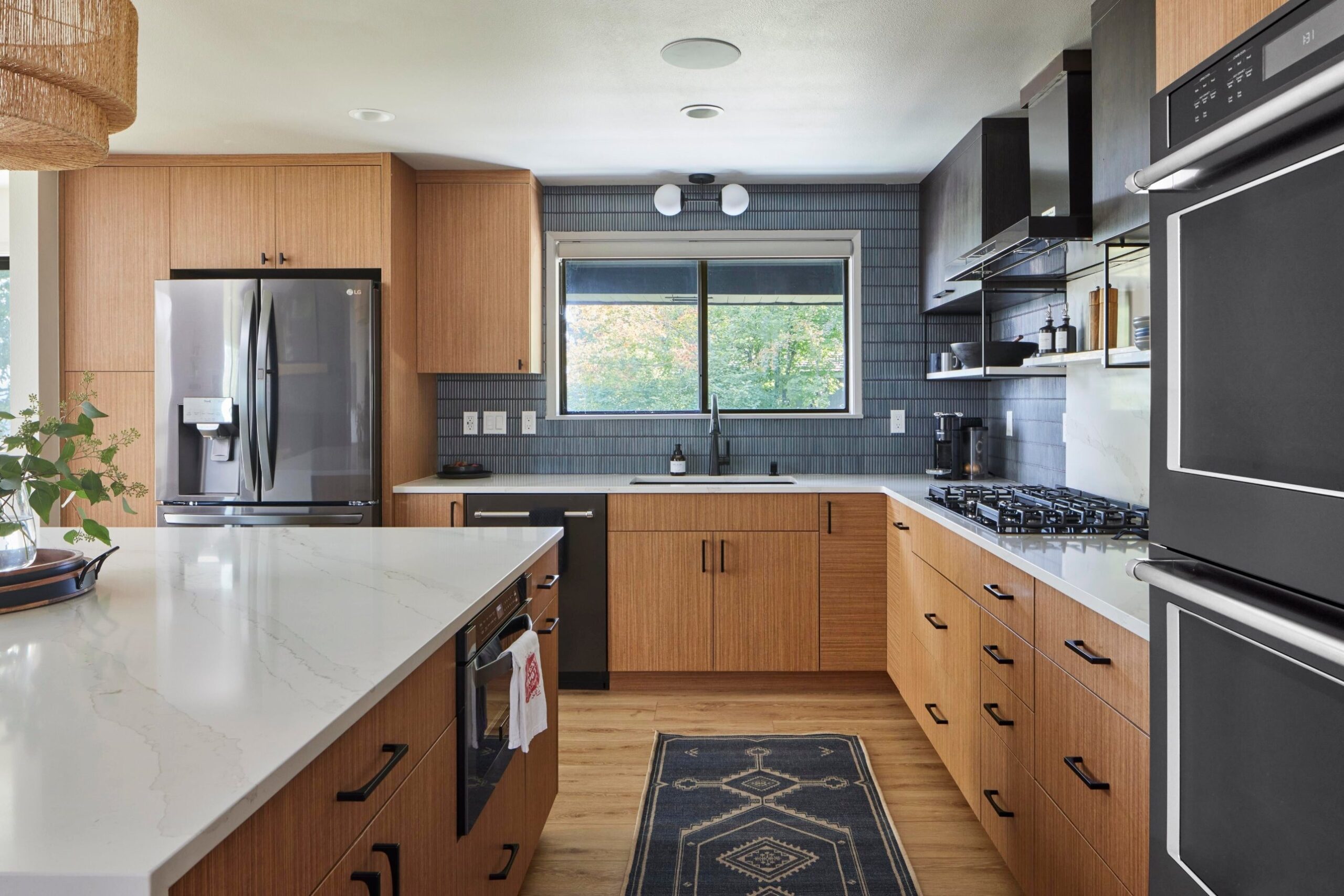 Kitchen view with a wide window over a black undermount sink, dark blue vertical tile wrapping the wall, a two-globe wall-mounted light fixture above the window, teak cabinets, and a dark blue patterned runner rug on the floor.