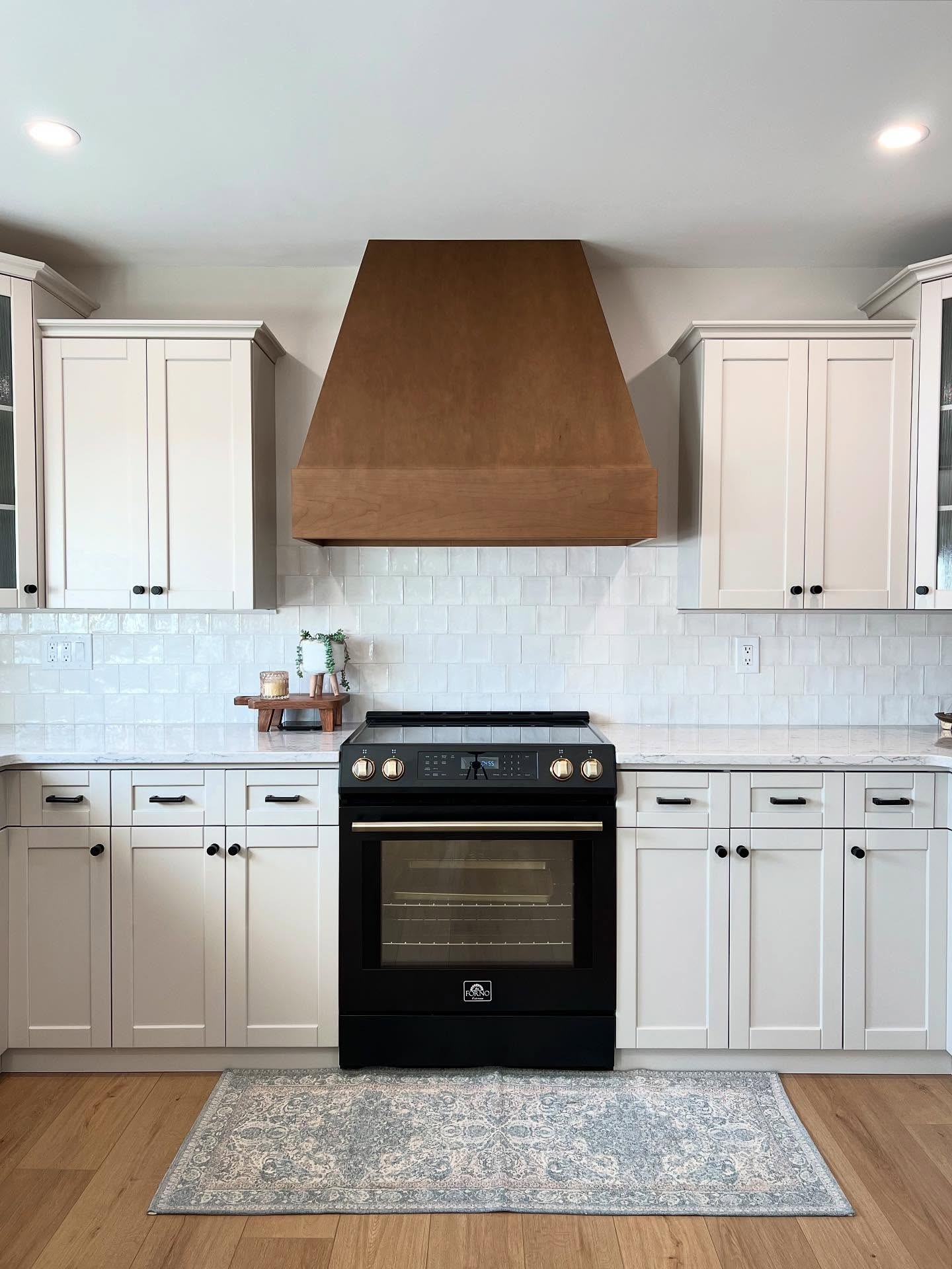 Straight-on view of the stove wall with a warm wood vent hood centered between soft greige Shaker upper cabinets, a white textured square tile backsplash, a matte black Forno slide-in range with brushed brass knobs, white quartz counters, and a blue vintage runner rug on light oak flooring.