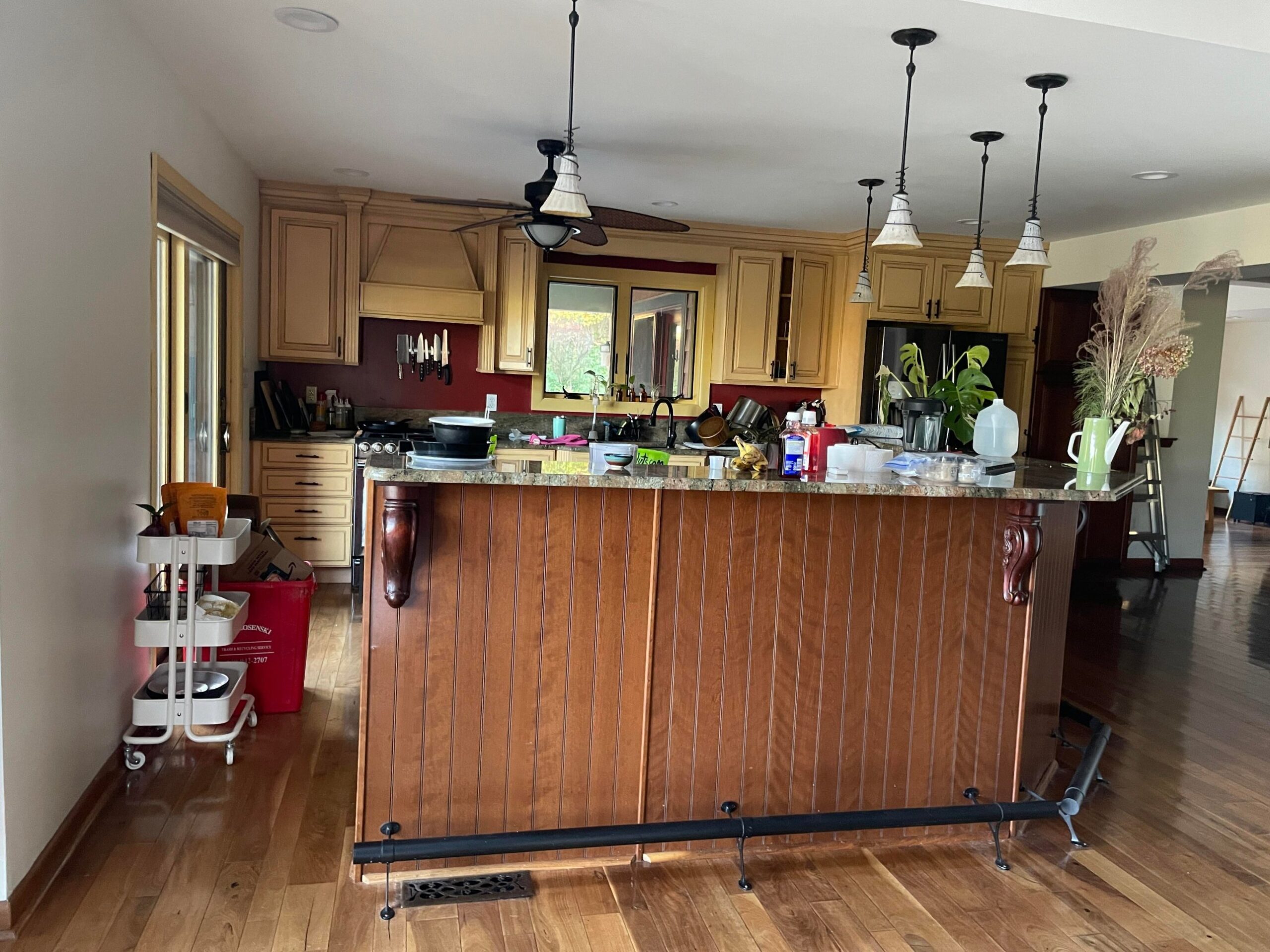 Wide view of a yellow glazed Tuscan kitchen with a cherry brown beadboard island, green granite counters, a tropical ceiling fan, globe pendant lights on wrought iron chains, and burgundy accent walls.