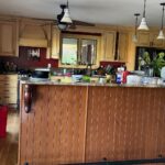 Wide-angle 16:9 view of a yellow glazed Tuscan kitchen with a cherry brown beadboard island, green granite counters, globe pendant lights, and burgundy accent walls before the Providence Blue paint makeover.