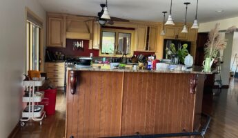 Wide-angle 16:9 view of a yellow glazed Tuscan kitchen with a cherry brown beadboard island, green granite counters, globe pendant lights, and burgundy accent walls before the Providence Blue paint makeover.