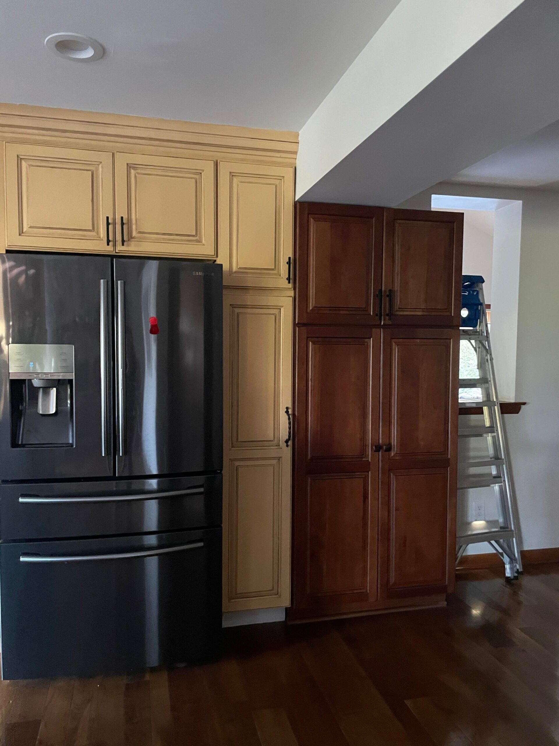 Yellow glazed cabinets above a black Samsung four-door fridge, with a tall cherry-stained pantry tower on the right and an aluminum ladder set up for painting.