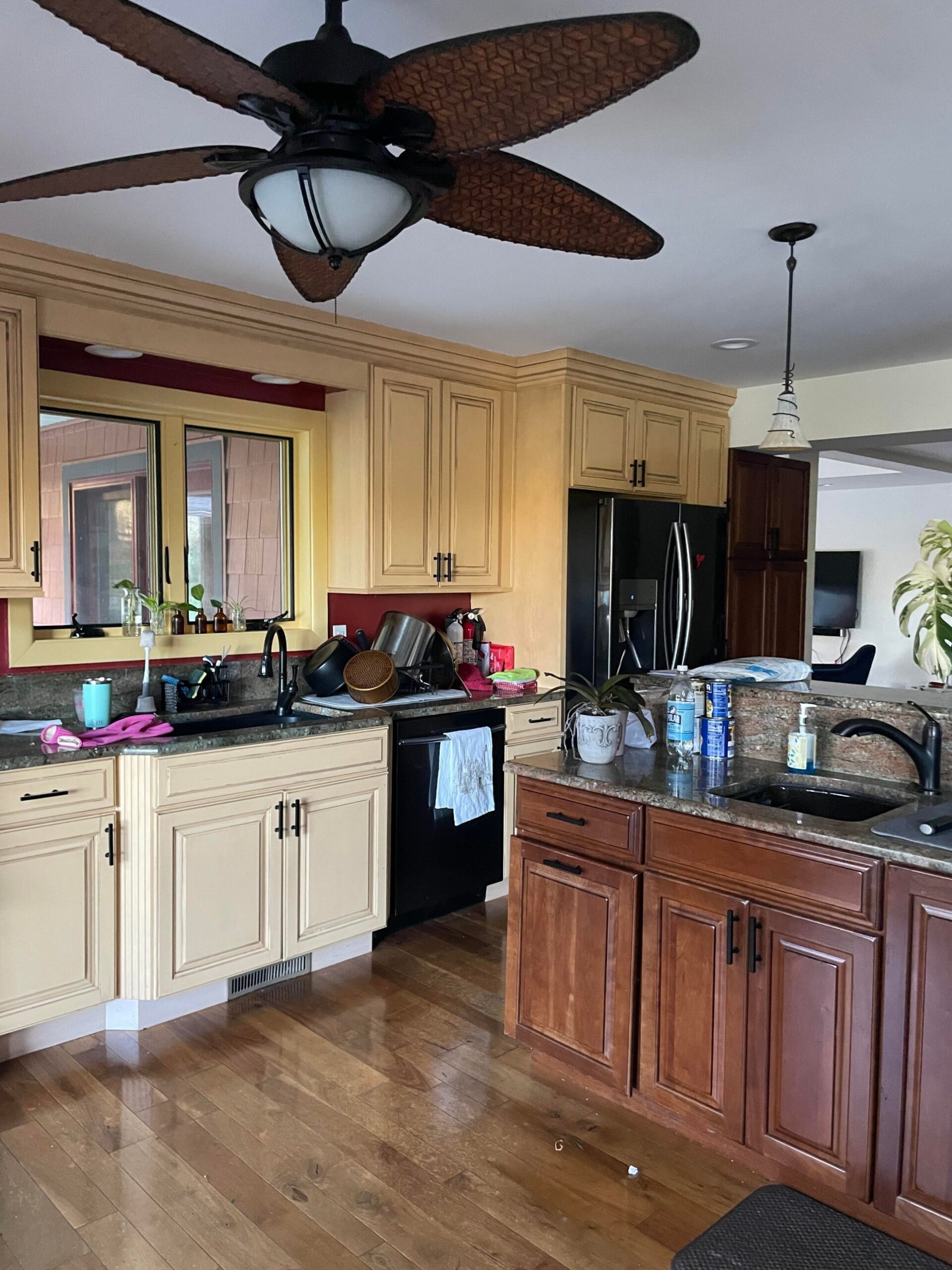 Yellow glazed lower cabinets with a black undermount sink and dishwasher, next to brown cherry cabinets on a secondary counter, under the tropical bamboo ceiling fan.