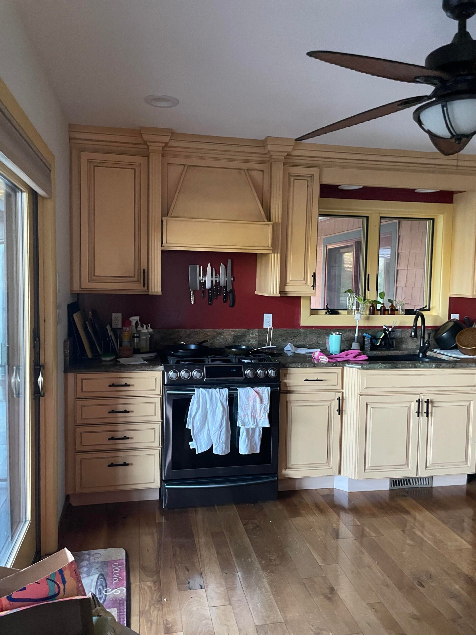 Yellow glazed cabinets around a black gas range with burgundy accent wall, magnetic knife strip, ornate hood with corbel detailing, and a window with yellow-painted trim.